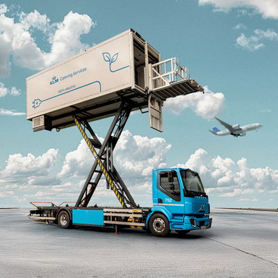 Blue electric catering Volvo truck with elevated platform labeled "KLM Catering Services," servicing planes under a clear sky.